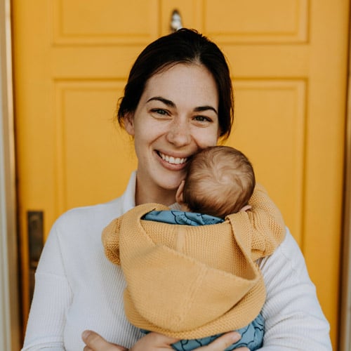 Mother holding new baby wrapped in a yellow shawl in front of a yellow door